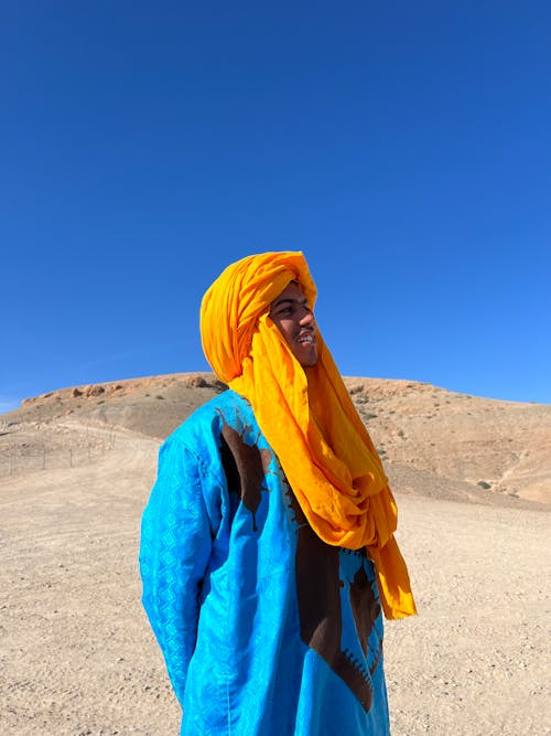 Free A man in vibrant traditional attire stands in a Moroccan desert landscape under a clear blue sky. Stock Photo