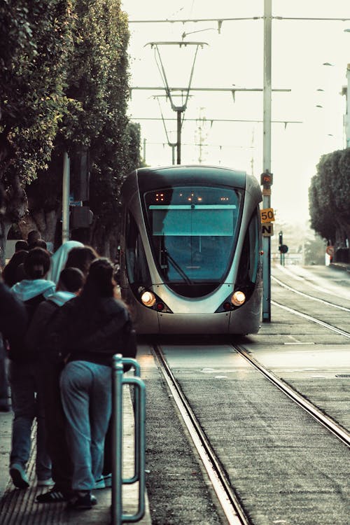 Free A sleek tram approaches an urban street station with people waiting on the side. Stock Photo