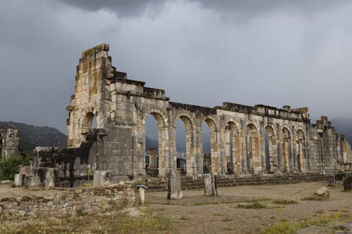 Free Ancient Roman ruins of Volubilis in Morocco, showcasing stone arches and dramatic cloudy sky. Stock Photo