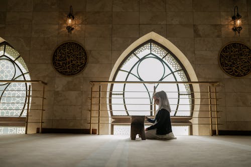 Free A Muslim girl in hijab sitting indoors, reading Quran during Ramadan. Peaceful religious moment. Stock Photo