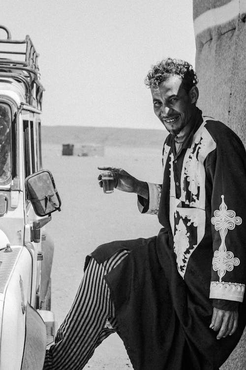 Free Man in traditional Moroccan attire enjoying tea by a car in Merzouga desert. Stock Photo