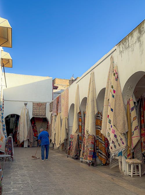 Free A picturesque market alley featuring traditional rugs and a person sweeping on a sunny day. Stock Photo