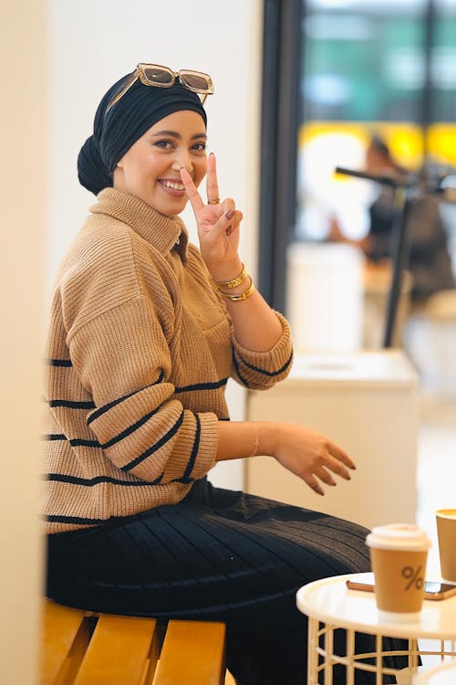 Free Smiling woman sitting at a café table with a peace sign and coffee cup, Dar-el-Beida. Stock Photo