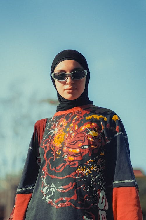 Free A stylish portrait of a modern hijabi in sunglasses and graphic tee against a blue sky in Kenitra, Morocco. Moroccan darija