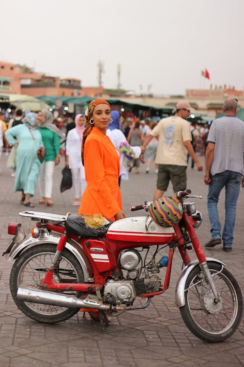 Free Woman in bright orange outfit posing with a vintage motorcycle in Marrakesh market. Stock Photo