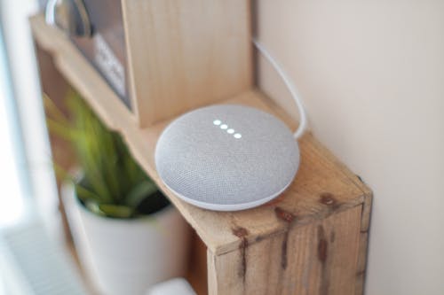 Free A Google Home Mini smart speaker on a wooden shelf, blending technology with home decor. Stock Photo