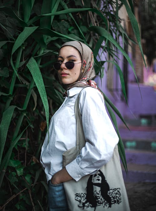 Free Fashionable woman in hijab and sunglasses poses outdoors with greenery backdrop. Stock Photo