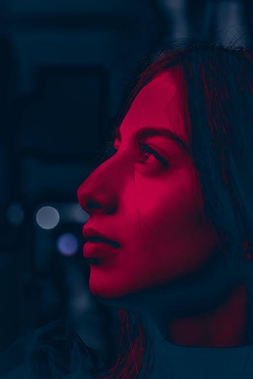 Free Striking portrait of a young woman in red-tinted light, capturing an elegant side profile with a city backdrop. Stock Photo